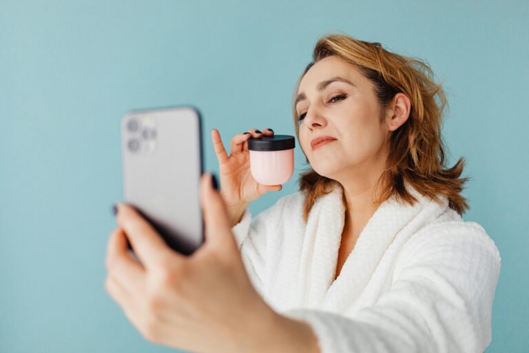 Woman in bathrobe taking a selfie holding a skincare product against a blue background.